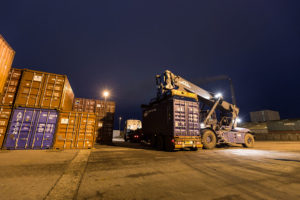 A large heavy-duty forklift preparing to lift a shipping container off a truck. Illustrating Multiterminal's expertise in container handling services, ensuring smooth loading, unloading, and transshipment across sea, rail, and road transport modes.